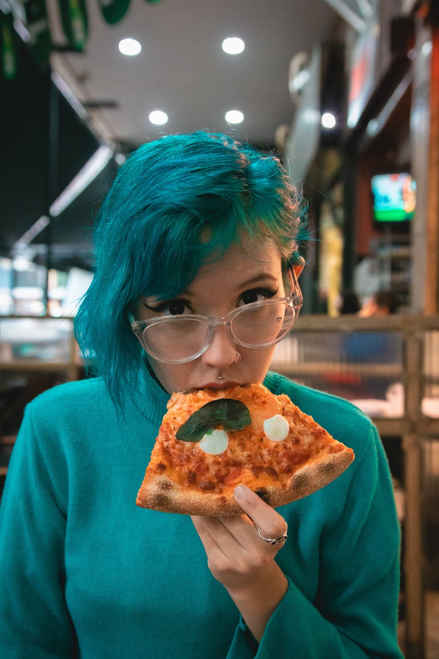 Femme aux cheveux bleus avec lunettes qui mange une pointe de pizza