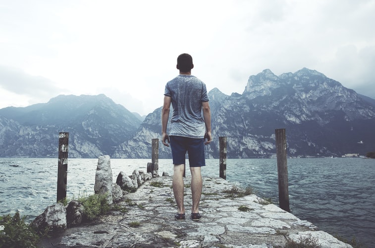 Homme debout sur un quai en béton gris face à un plan d'eau et des montagnes dans la journée